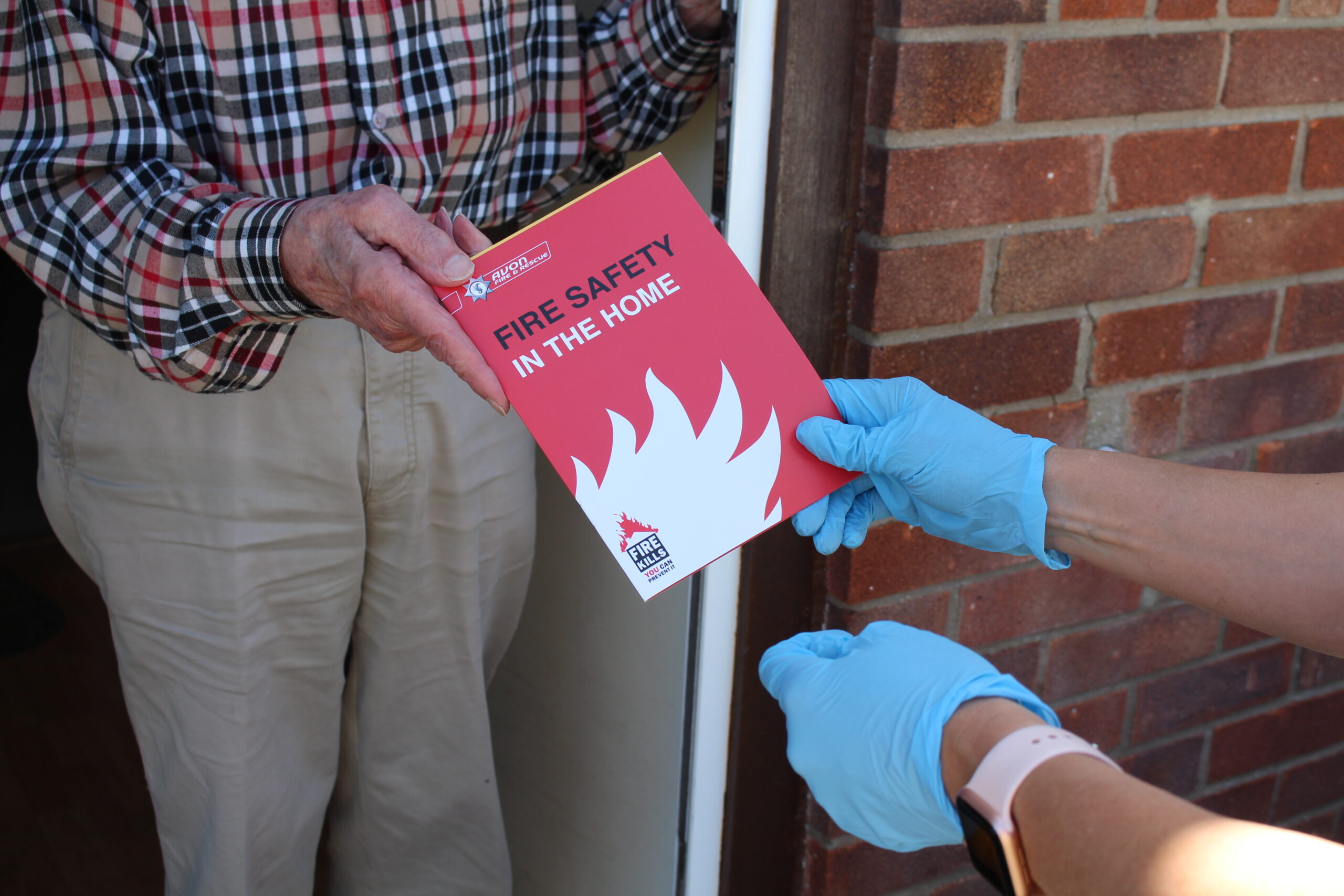 gloved hands giving red and white home fire safety leaflet to elderly man at doorstep.