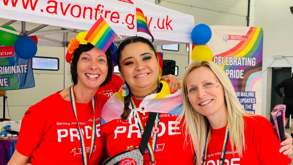 Image of three women donning red shirts and waving rainbow flags.