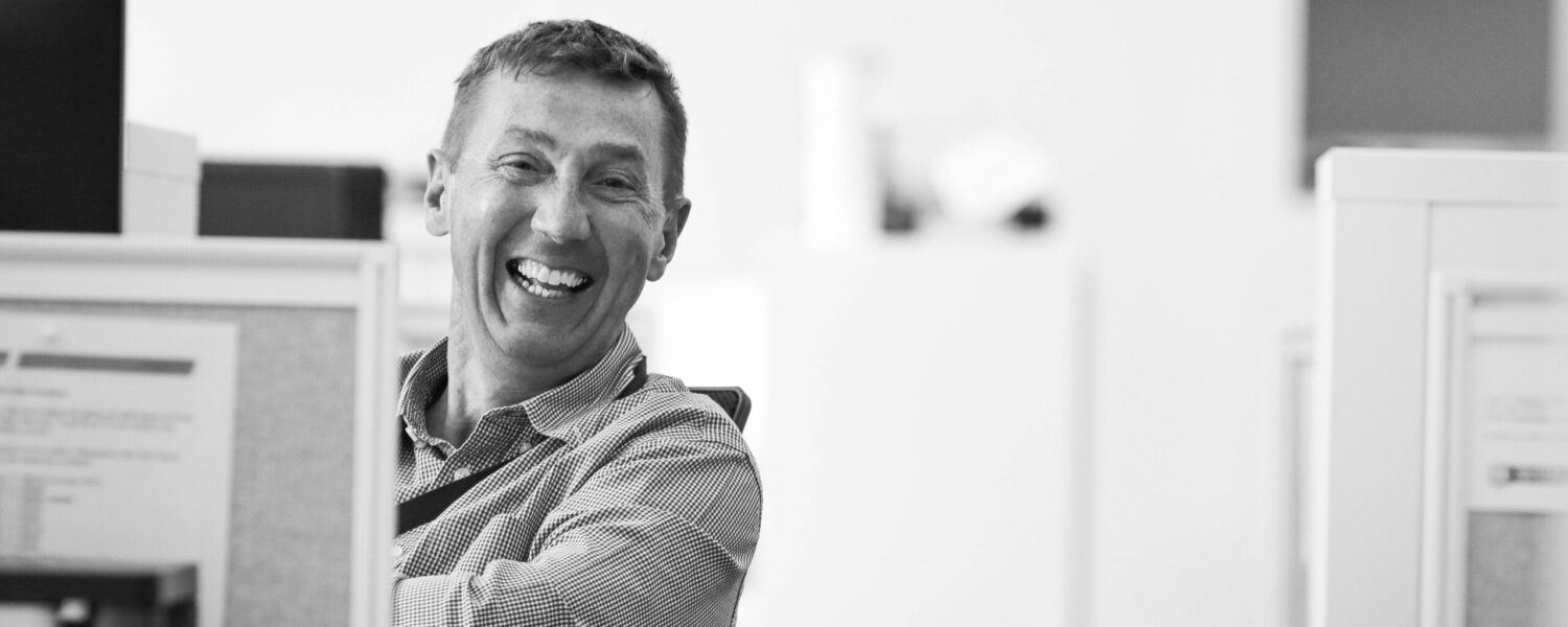 male corporate staff member sitting at screen smiling at photographer