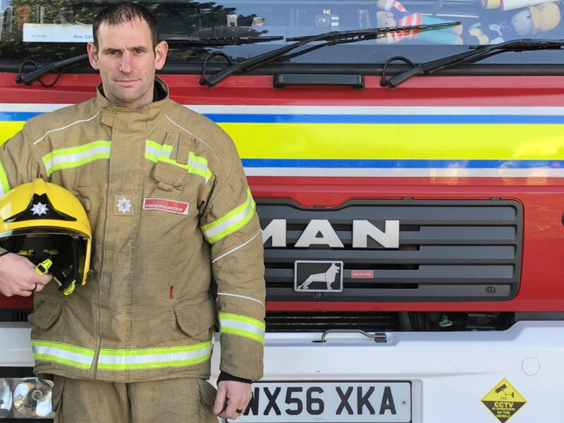 white male firefighter in full kit, holding helmet to his side. In front of fire engine