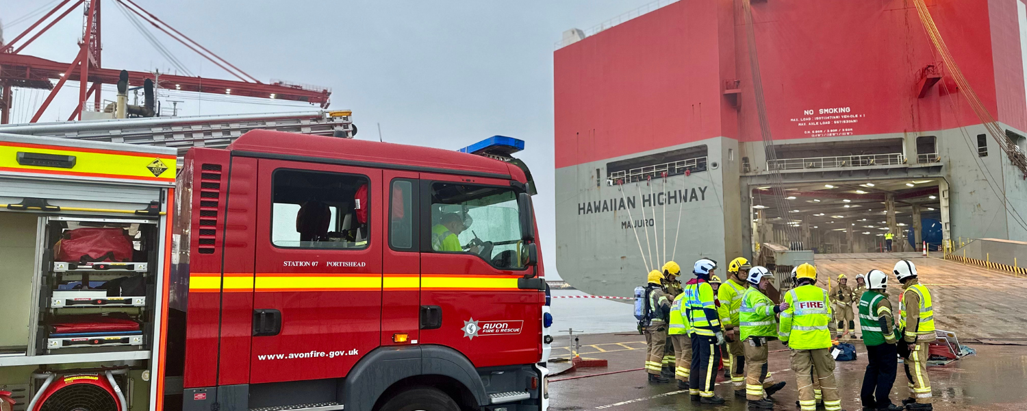 large cargo ship on the right with a fire crew in front of it. fire engine parked on the left.