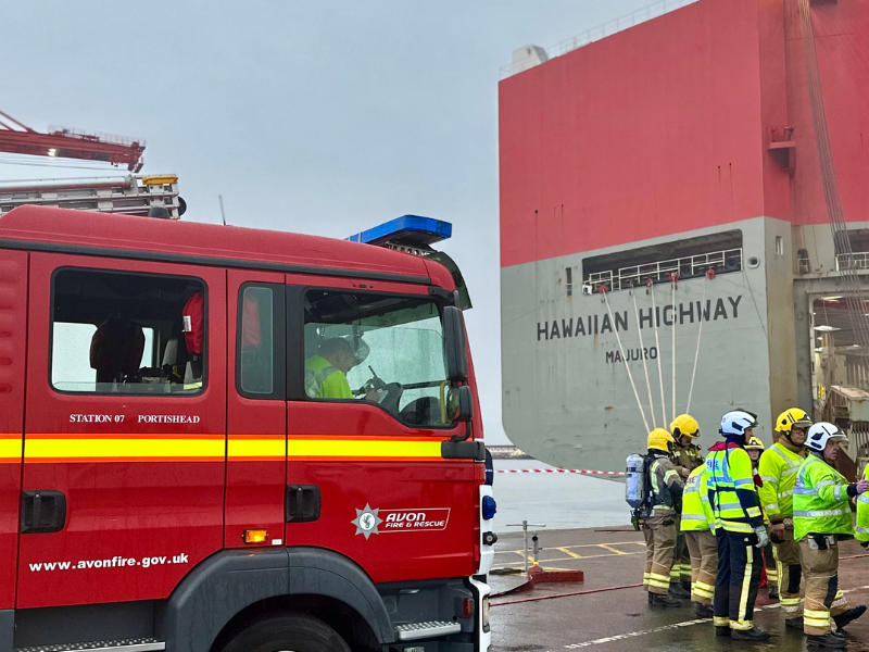 large cargo ship on the right with a fire crew in front of it. fire engine parked on the left.
