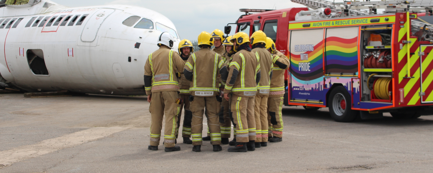 photo showing a group of firefighters in the centre standing in front of a crashed plane (left) and a fire engine (right) with a rainbow design on it