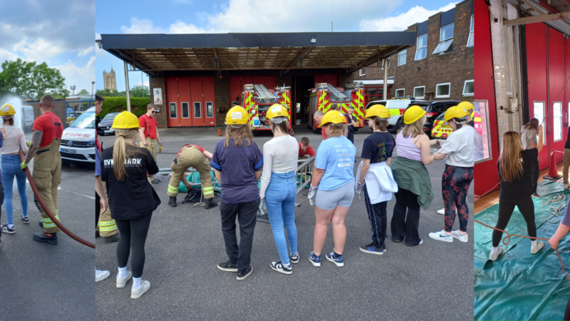 group of college students wearing casual clothes and firefighter helmets interacting with uniformed firefighters at Yate Fire Station, within the station yard