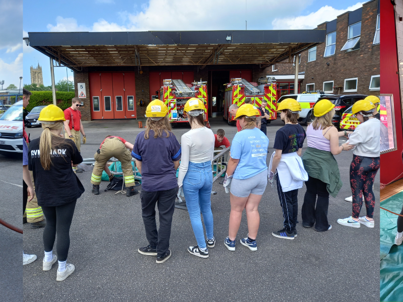 group of college students wearing casual clothes and firefighter helmets interacting with uniformed firefighters at Yate Fire Station, within the station yard