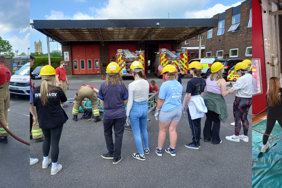 group of college students wearing casual clothes and firefighter helmets interacting with uniformed firefighters at Yate Fire Station, within the station yard