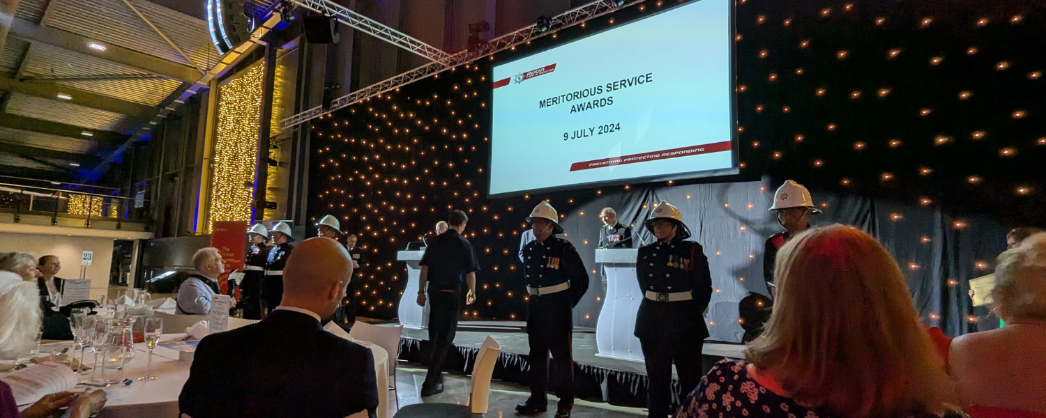 A group of uniformed individuals stands in front of a stage, while an audience watches. The screen on stage reads 