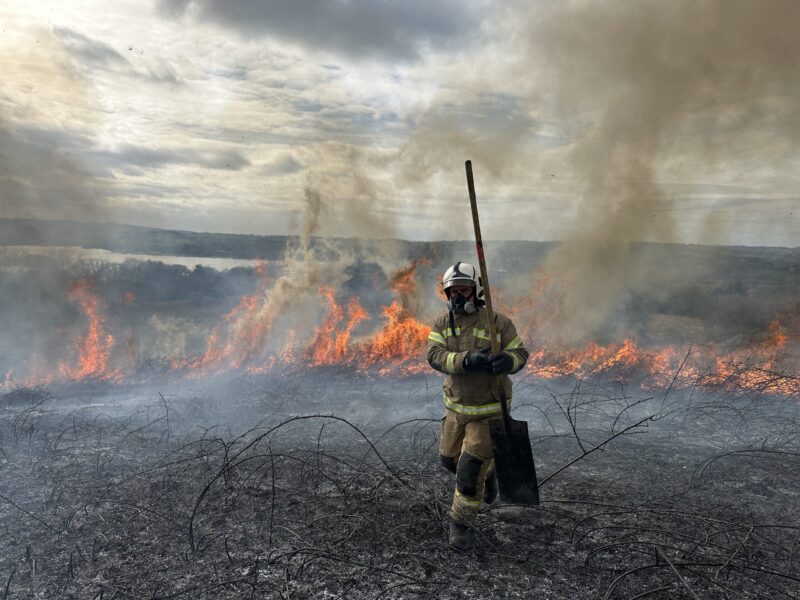 Large grass fire in background, with firefighter in foreground seen fighting the fire