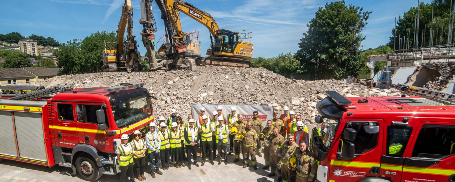 Staff from Avon Fire & Rescue Service and representatives from Willmott Dixon stand between two fire engines in front of the demolished fire station at Bath.