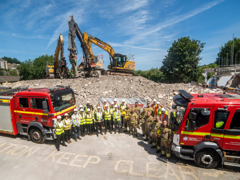 Staff from Avon Fire & Rescue Service and representatives from Willmott Dixon stand between two fire engines in front of the demolished fire station at Bath.