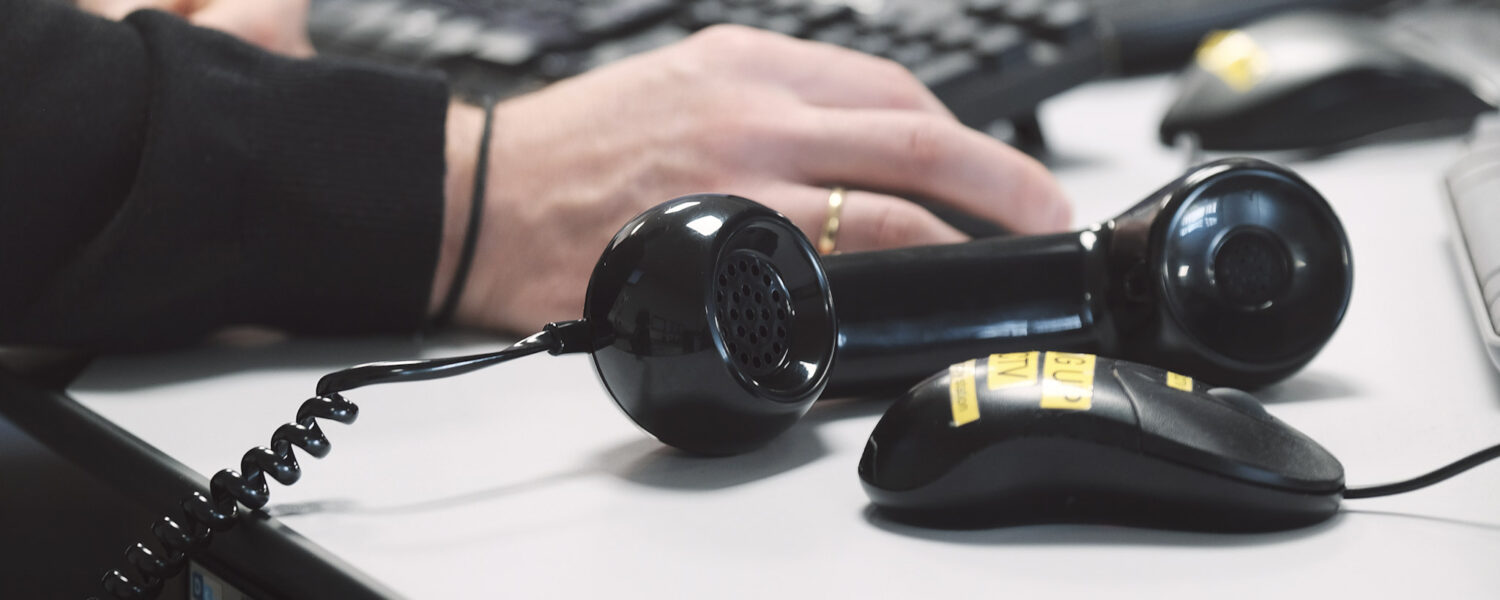 black handset of a landline phone lying on its side next to a computer mouse on a desk with someone using a keyboard behind
