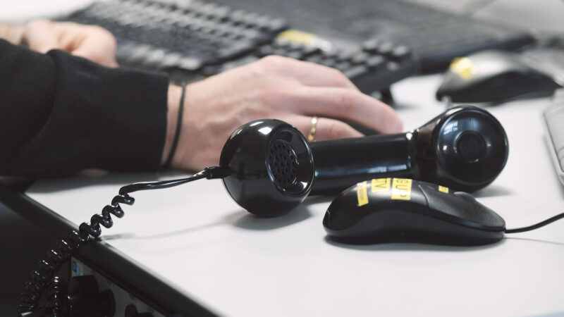 black handset of a landline phone lying on its side next to a computer mouse on a desk with someone using a keyboard behind