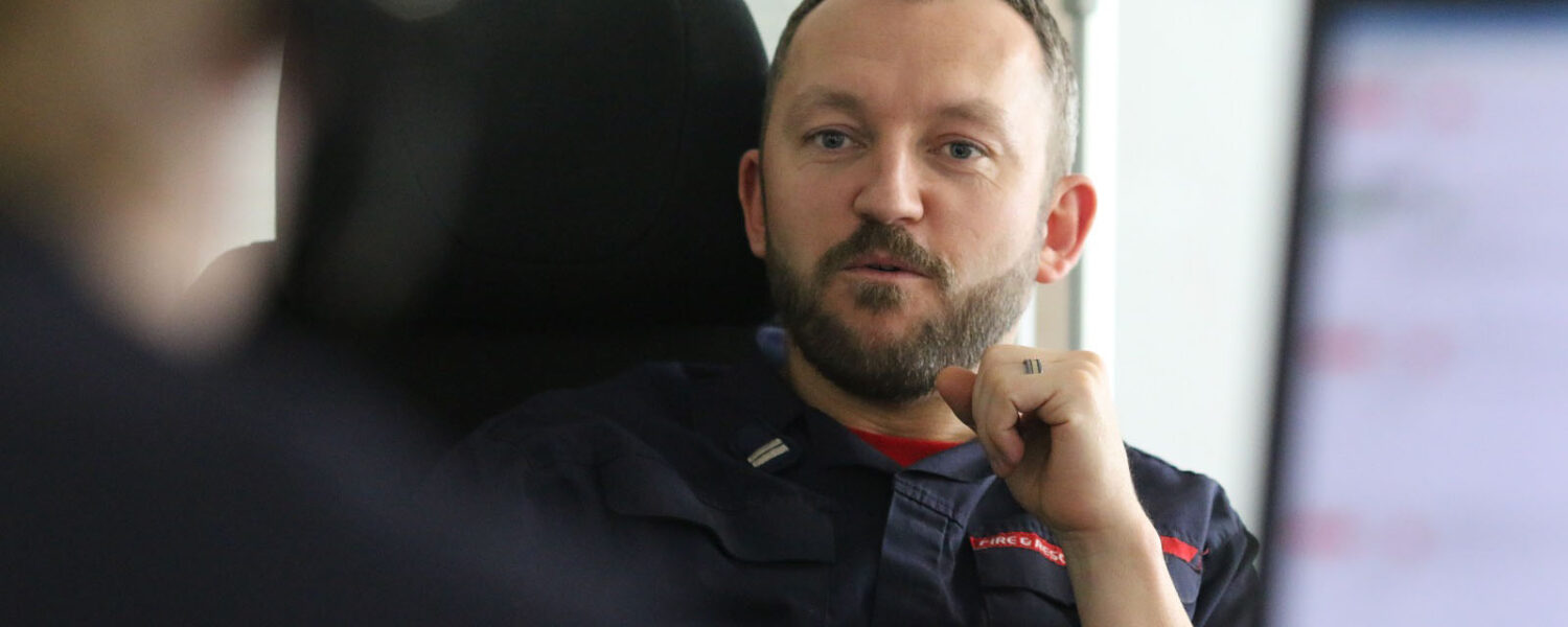male control firefighter in blue shirt uniform sitting talking to another control firefighter with a computer screen