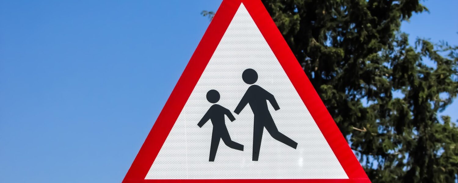 a red triangular road sign featuring an icon of two children in front of a blue sky and tall cypress tree