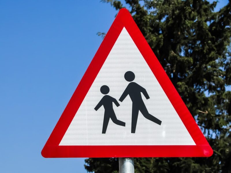 a red triangular road sign featuring an icon of two children in front of a blue sky and tall cypress tree