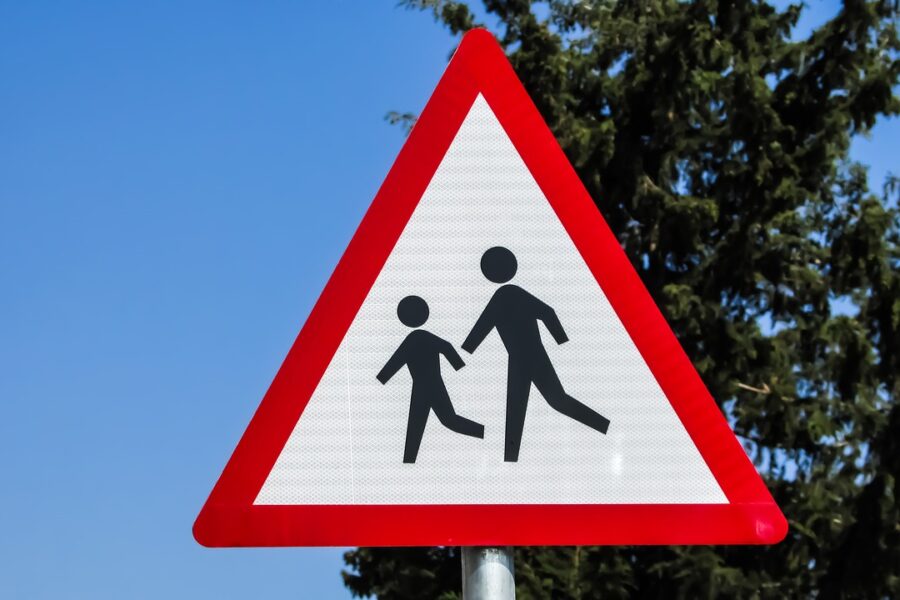 a red triangular road sign featuring an icon of two children in front of a blue sky and tall cypress tree