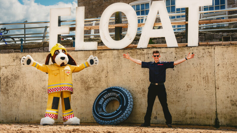 fido the firedog large dog mascot standing next to a firefighter both doing the 'float to live' position by stretching out their arms and legs. They are on a beach, with the word 'float' behind them.