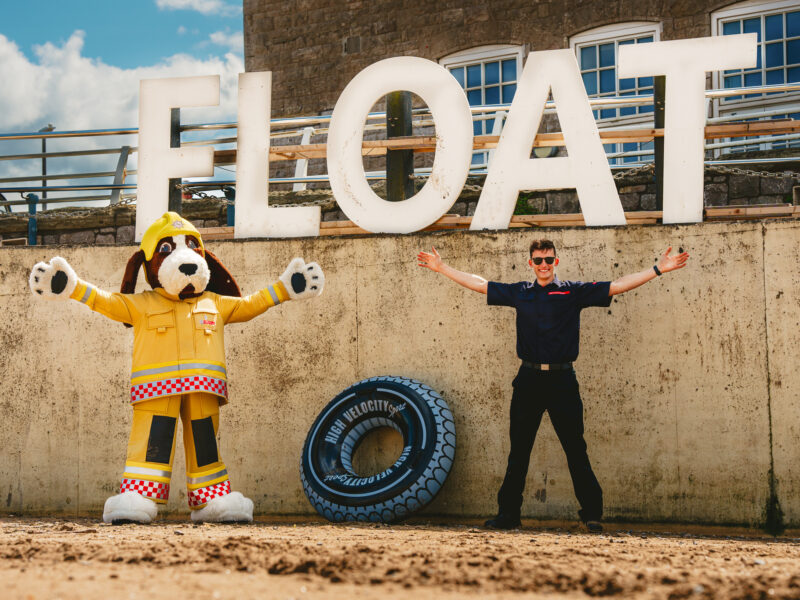 fido the firedog large dog mascot standing next to a firefighter both doing the 'float to live' position by stretching out their arms and legs. They are on a beach, with the word 'float' behind them.
