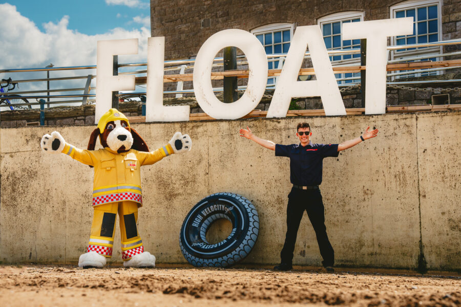 fido the firedog large dog mascot standing next to a firefighter both doing the 'float to live' position by stretching out their arms and legs. They are on a beach, with the word 'float' behind them.