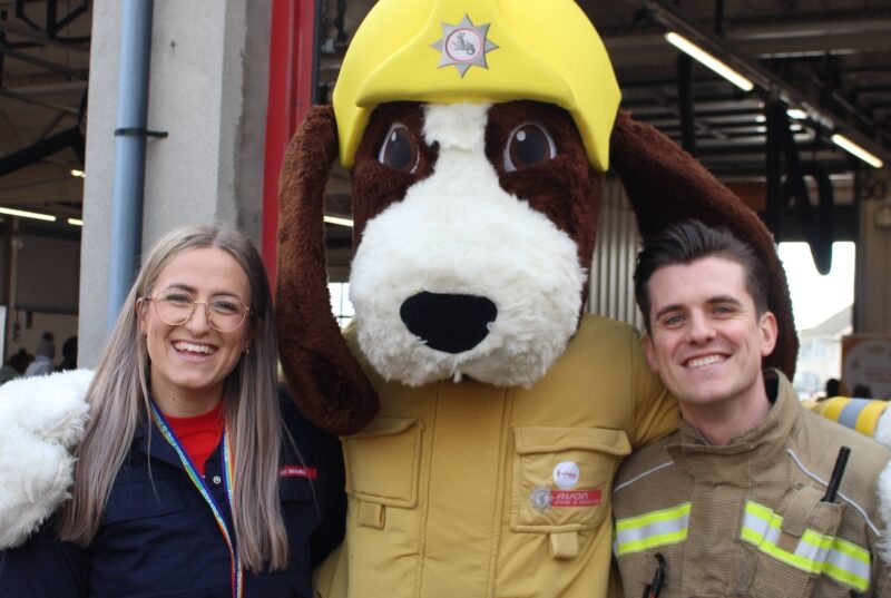 smiling blonde woman in glasses and red and blue uniform with Fido the fire dog mascot and male firefighter with brown hair