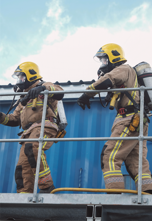 Two firefighters walking to the left wearing full fire kit and breathing apparatus in front of blue metal sheeting with blue sky above