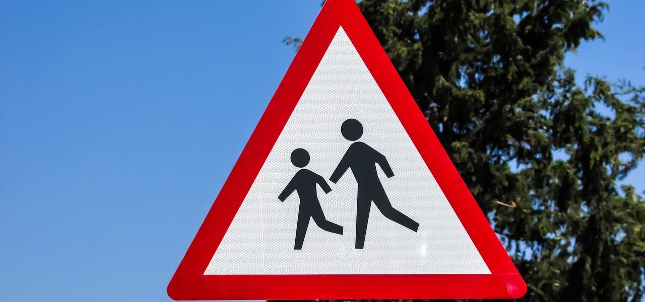 a red triangular road sign featuring an icon of two children in front of a blue sky and tall cypress tree